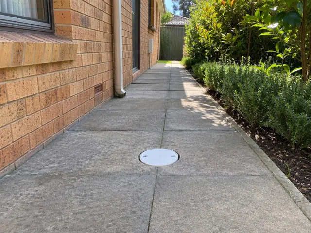 Dry paved side path with a round drain cover beside a brick house and garden in Mount Colah