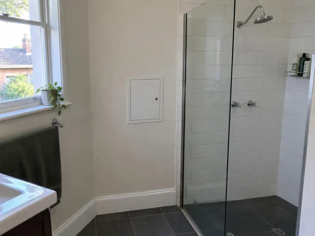 Small access panel on a painted bathroom wall beside a tiled shower and window in Waitara